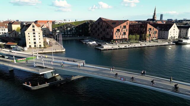 Aerial view of a modern bridge spanning dark waters, connecting red brick buildings under a bright sky, a blend of urban design, Copenhagen, Denmark.