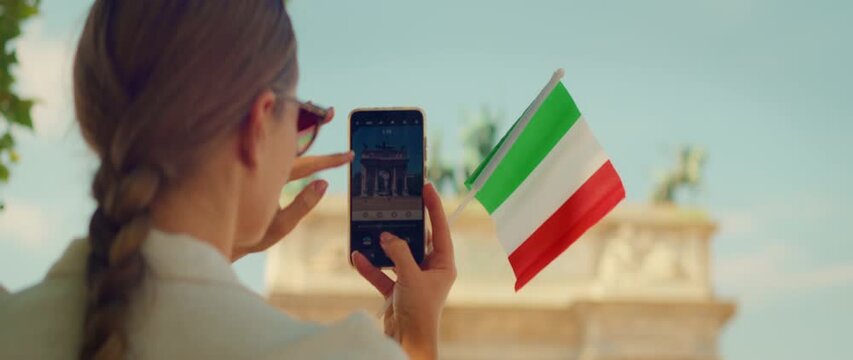翻译标题：在米兰，一名游客手持意大利国旗用手机拍摄纪念碑